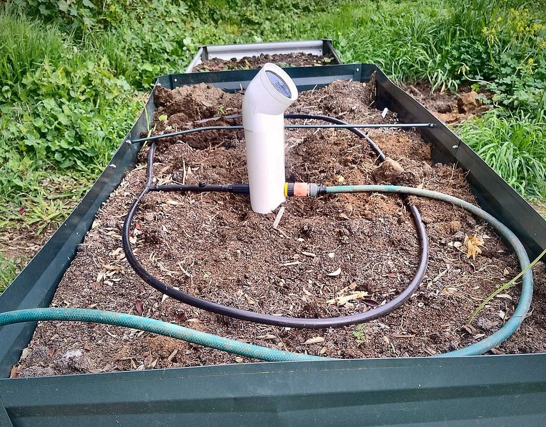 A TSAM Sundew unit installed in the middle of a raised vegetable bed, with drip line looped around it and a garden hose connected to its inlet.